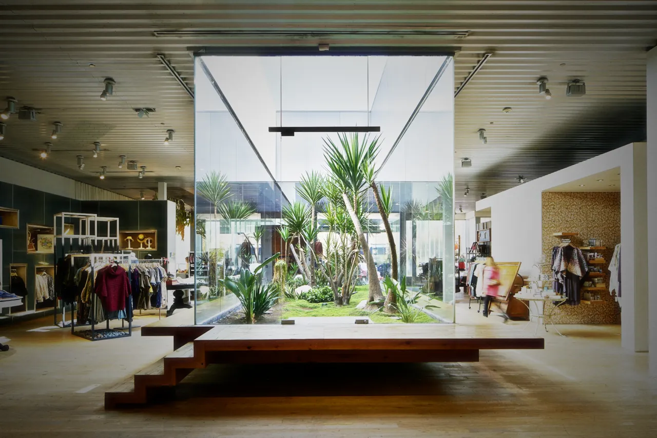 Anthropologie Dos Lagos: a glass room of yucca and grass open to the sky, behind a timber plinth, with the clothes around it