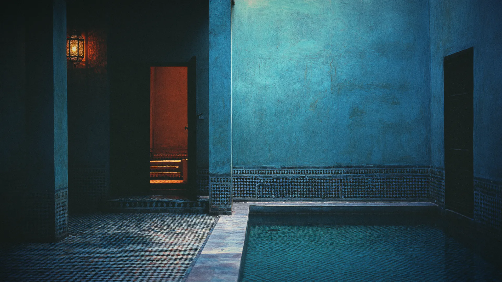 A blue courtyard at night, one warm lantern glowing in a doorway beyond the pool