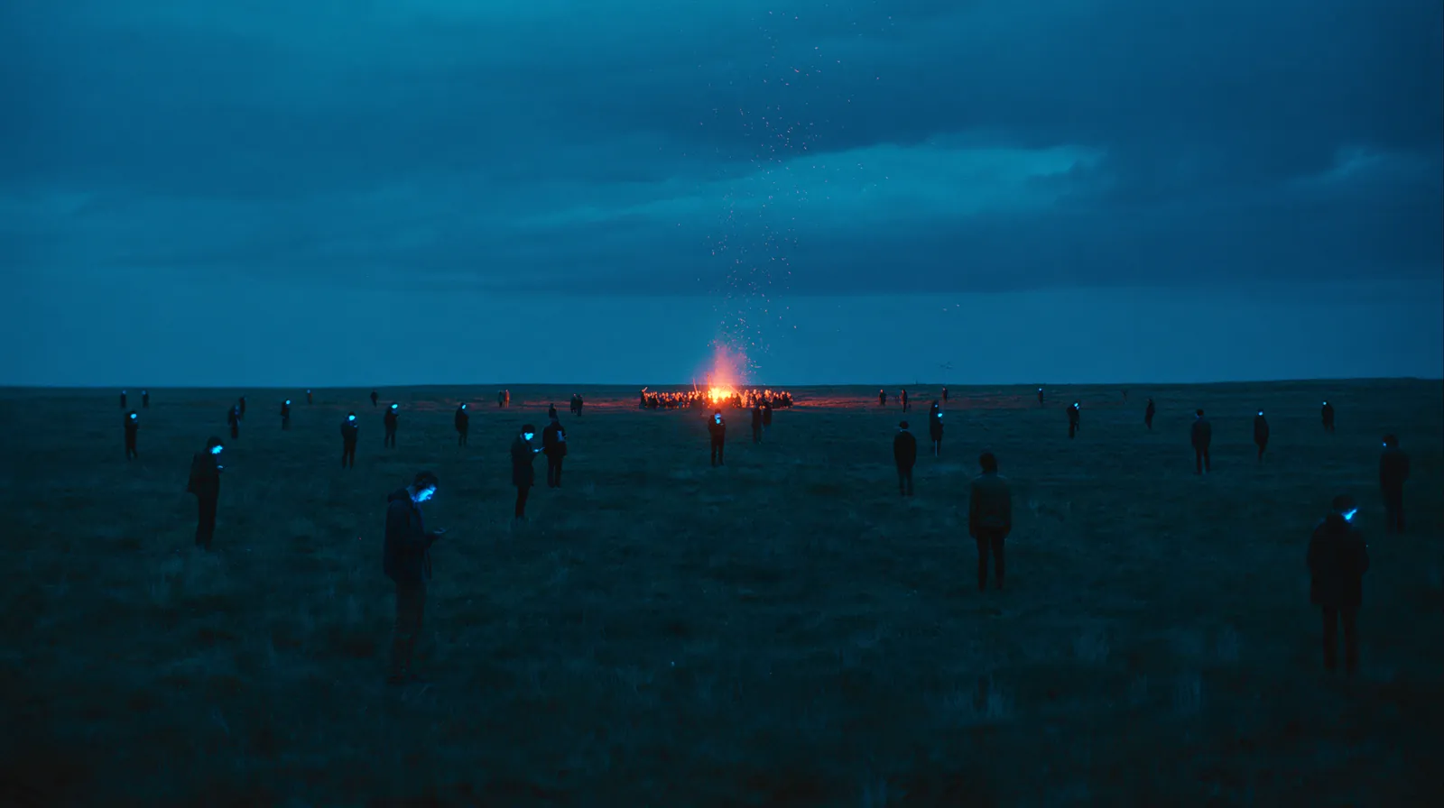 People standing alone on a dark plain, each lit by a phone, and one fire in the distance with a crowd around it